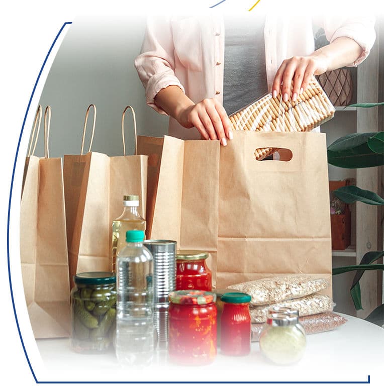 Hands packing jars, canned goods, and crackers into brown paper grocery bags for food donation.